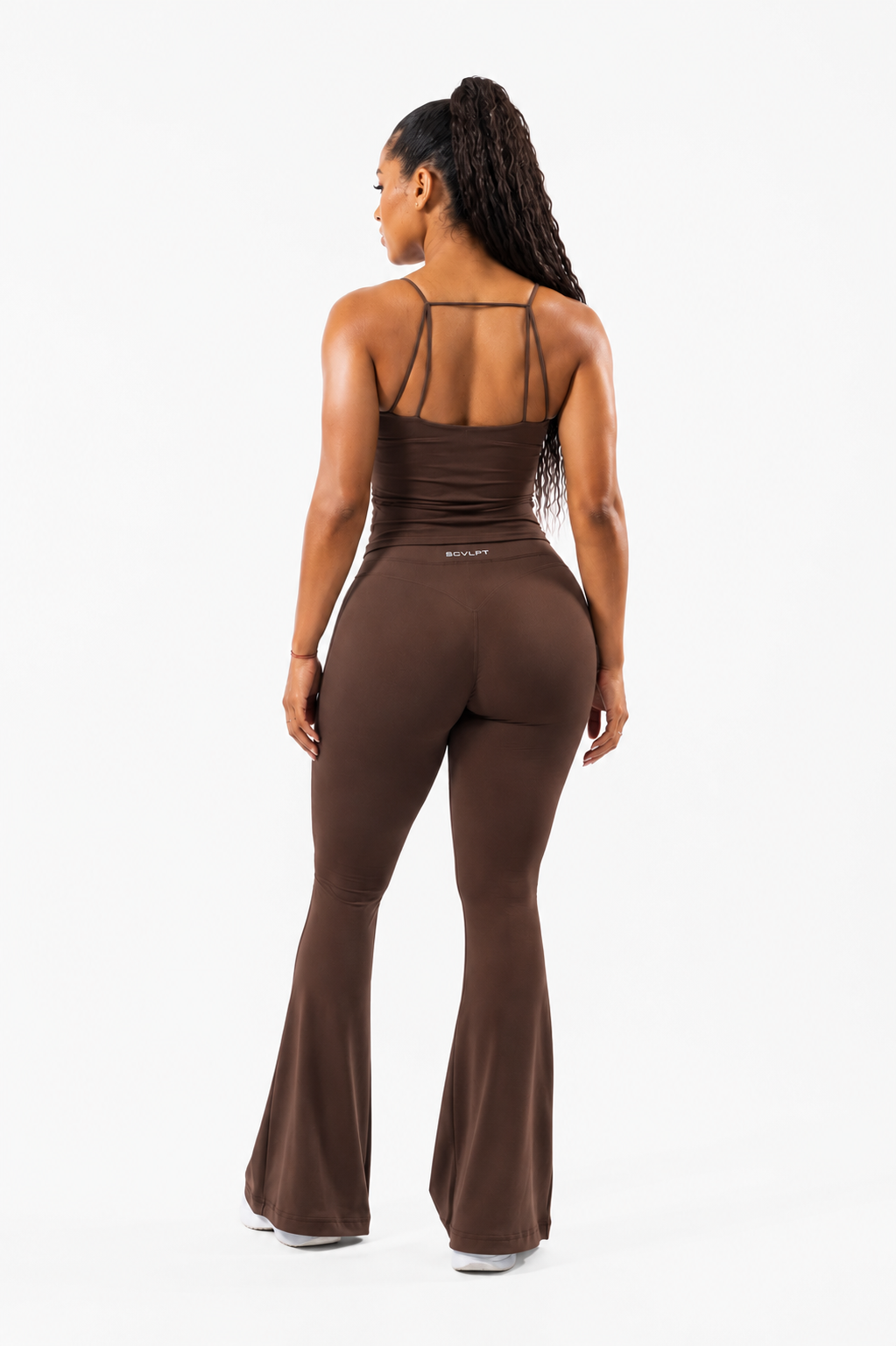 A woman modeling a brown sleeveless top with thin straps and flared leggings, standing against a plain white background.