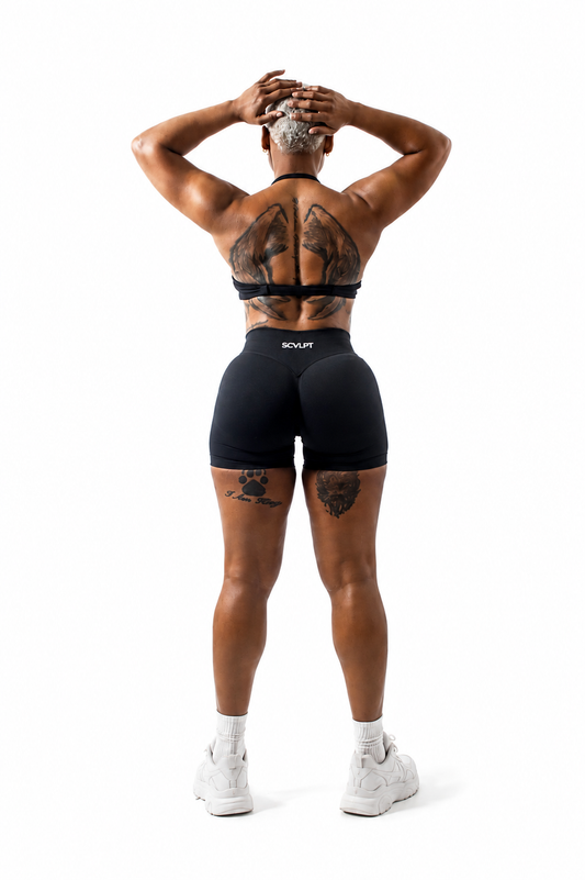 A woman is shown from behind wearing black athletic shorts and a black sports bra, with white sneakers, against a solid white background.