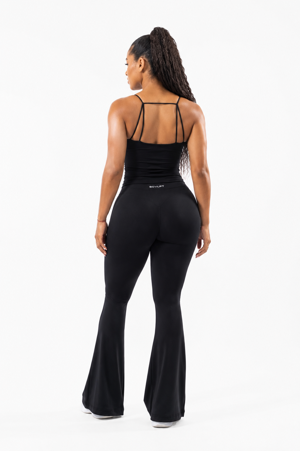 A woman shown from the back wearing a black sleeveless top with thin shoulder straps and flared leggings, photographed against a clean white studio background.
