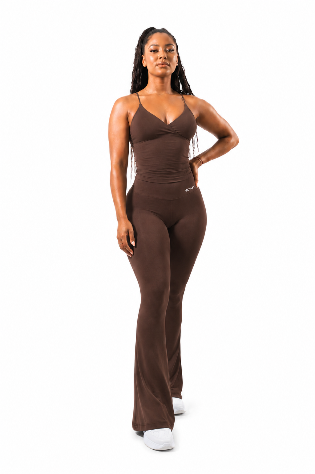 A woman modeling brown flared leggings with a sleeveless strappy top standing against a plain white background.