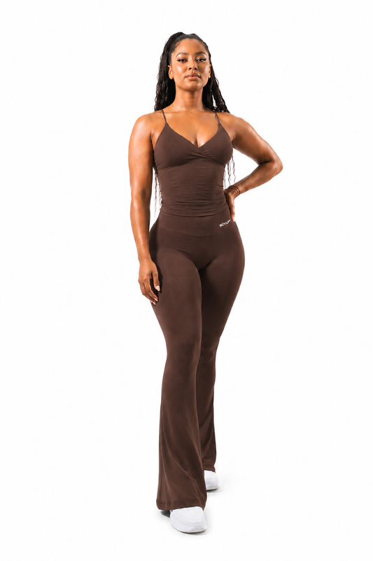 A woman modeling brown flared leggings with a sleeveless strappy top standing against a plain white background.