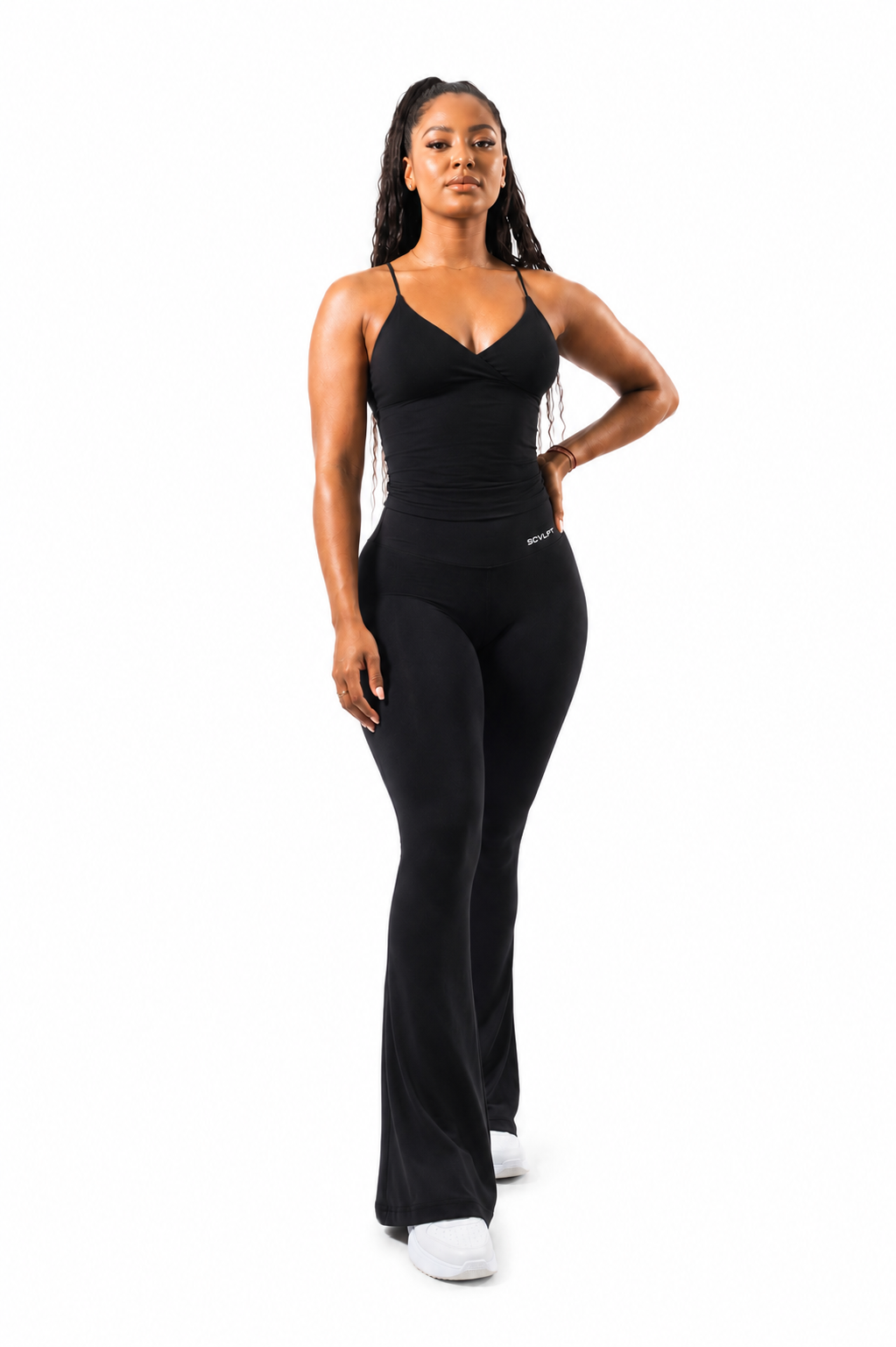 A woman modeling brown flared leggings with a sleeveless strappy top standing against a plain white background.
